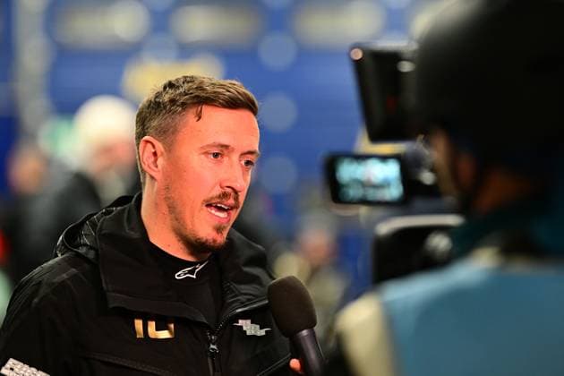 Max Kruse in schwarzer Alpinestars-Jacke mit der Nummer 10 gibt einem TV-Team ein Interview. Im Hintergrund ist ein blauer Sponsoren-Backdrop zu sehen. Der Kameramann hält die Kamera auf den Fahrer gerichtet, während ein Mikrofon ins Bild ragt. Die Szene findet im Fahrerlager des Nürburgrings statt. Fahrer-Interview beim 24h Nürburgring 2026 im Fahrerlager Copyright: xMichaelxGelhardtx