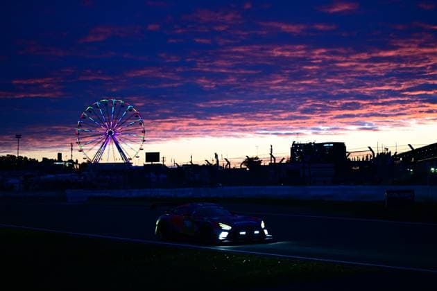 Ein GT3-Rennfahrzeug durchquert bei Sonnenuntergang die Strecke am Nürburgring. Die Scheinwerfer des Fahrzeugs leuchten in der Abenddämmerung, während am Horizont ein bunt beleuchtetes Riesenrad und die Silhouette der Fahrerlager-Infrastruktur zu sehen sind. Der dramatisch gefärbte Himmel mit orangefarbenen und violetten Wolken verleiht der Szene eine besondere Atmosphäre. GT3-Fahrzeug bei Sonnenuntergang am Nürburgring mit Riesenrad Copyright: xMichaelxGelhardtx