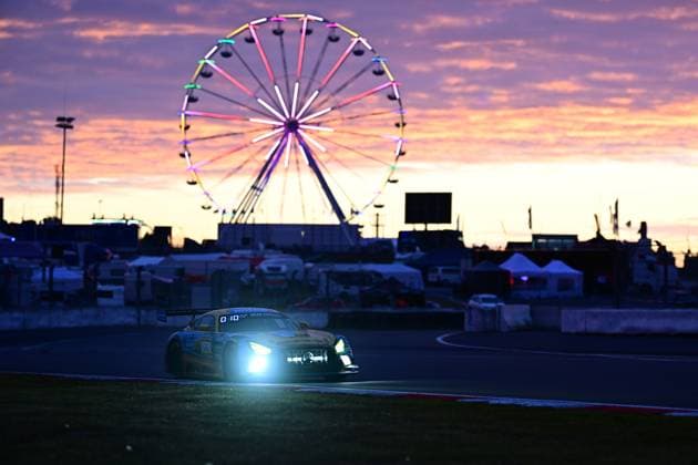 Der Mercedes-AMG GT3 Adam Christodoulou & Christopher Lulham & Marek Böckmann & Mikaël Grenier (PROsport Racing) von PROsport Racing durchfährt in der Abenddämmerung die Boxengasse des Nürburgrings. Die Scheinwerfer leuchten hell in der hereinbrechenden Nacht, während im Hintergrund das farbig beleuchtete Riesenrad und der dramatische Sonnenuntergang mit violett-orangenem Himmel eine eindrucksvolle Kulisse bilden. Das Bild vermittelt die besondere Atmosphäre des 24-Stunden-Rennens. Mercedes-AMG GT3 26 bei Sonnenuntergang am Nürburgring Copyright: xMichaelxGelhardtx