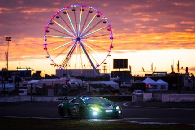 Der Lamborghini Huracán GT3 Marco Mapelli & Nicholas Yelloly & Nicky Catsburg (Red Bull Team ABT) von Red Bull Team ABT fährt im Dämmerungslicht über die Strecke am Nürburgring. Die eingeschalteten Scheinwerfer leuchten hell, während im Hintergrund ein farbig beleuchtetes Riesenrad vor einem spektakulären orangefarbenen Sonnenuntergangshimmel erstrahlt. Die Atmosphäre vermittelt die besondere Stimmung des 24-Stunden-Rennens beim Übergang von Tag zu Nacht. Lamborghini 130 von Red Bull Team ABT bei Sonnenuntergang am Nürburgring Copyright: xMichaelxGelhardtx