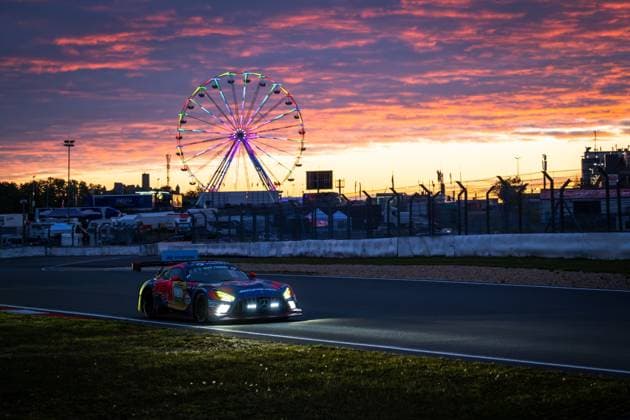 Der Mercedes-AMG GT3 Daniel Juncadella & Jules Gounon & Lucas Auer & Max Verstappen (Mercedes-AMG Team Verstappen Racing) von Mercedes-AMG Team Verstappen Racing durchfährt in der Abenddämmerung den Nürburgring. Die eingeschalteten Scheinwerfer leuchten die Strecke aus, während ein dramatisch rot-orangefarbener Sonnenuntergang den Himmel einfärbt. Im Hintergrund dreht sich das beleuchtete Riesenrad des Festgeländes, das die besondere Atmosphäre des 24-Stunden-Rennens unterstreicht. Das Red-Bull-Livery des Fahrzeugs ist im letzten Tageslicht gut erkennbar. Mercedes-AMG GT3 3 von Verstappen Racing beim Sonnenuntergang am Nürburgring Copyright: xMichaelxGelhardtx