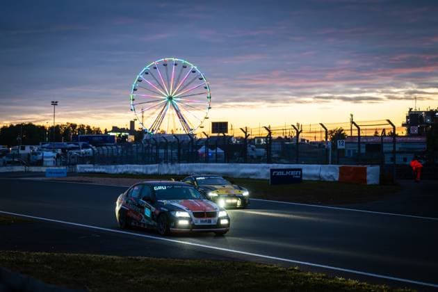 Der BMW 3er von EiFelkind Racing (Christopher Gruber & Henning Hausmeier & Markus Fischer & Michael Fischer & Oliver Frisse (Pistorius by EiFelkind Racing)) rollt in der Abenddämmerung über den Grand-Prix-Kurs des Nürburgrings. Das Fahrzeug mit der Startnummer 100 trägt die markante orange-schwarz-weiße Lackierung mit GRU Gran Turismo- und H&R-Sponsoring. Dahinter folgt ein weiteres Fahrzeug im Abblendlicht. Im Hintergrund leuchtet das beleuchtete Riesenrad des Fahrerlagers bunt vor einem dramatischen Sonnenuntergangshimmel in Orange und Violett. Die Streckenbeschilderung mit Falken Tyres und das Hyundai-Banner sind erkennbar. BMW 100 von EiFelkind Racing bei Sonnenuntergang am Nürburgring Copyright: xMichaelxGelhardtx