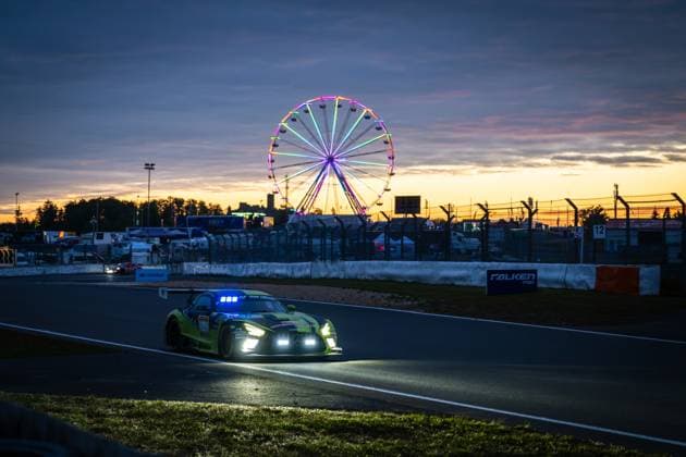 Ein neongelb-schwarzer Mercedes-AMG GT3 durchfährt in der Abenddämmerung eine Kurve am Nürburgring. Die eingeschalteten Scheinwerfer und die blauen Dachlichter leuchten kräftig in der hereinbrechenden Dunkelheit. Im Hintergrund erstrahlt ein bunt beleuchtetes Riesenrad vor einem dramatischen Sonnenuntergangshimmel in Orange- und Blautönen. Ein Falken Tyres-Banner ist an der Streckenbegrenzung sichtbar. Die Startnummer ist im Gegenlicht und durch die Bewegungsunschärfe nicht eindeutig lesbar. Mercedes-AMG GT3 im Sonnenuntergang am Nürburgring mit Riesenrad Copyright: xMichaelxGelhardtx