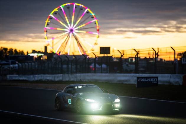 Der Toyota GR Supra 524; TSV 1860 Mühldorf von SRS Team Sorg Rennsport fährt in der Abenddämmerung auf dem Nürburgring. Die eingeschalteten Scheinwerfer leuchten hell in der einsetzenden Dunkelheit, während im Hintergrund das beleuchtete Riesenrad des Fahrerlagers in bunten Farben erstrahlt. Der goldene Sonnenuntergang verleiht der Szene eine stimmungsvolle Atmosphäre. Ein Falken Tyres-Banner ist an der Streckenbegrenzung zu sehen. Toyota GR Supra 524 bei Sonnenuntergang am Nürburgring Copyright: xMichaelxGelhardtx