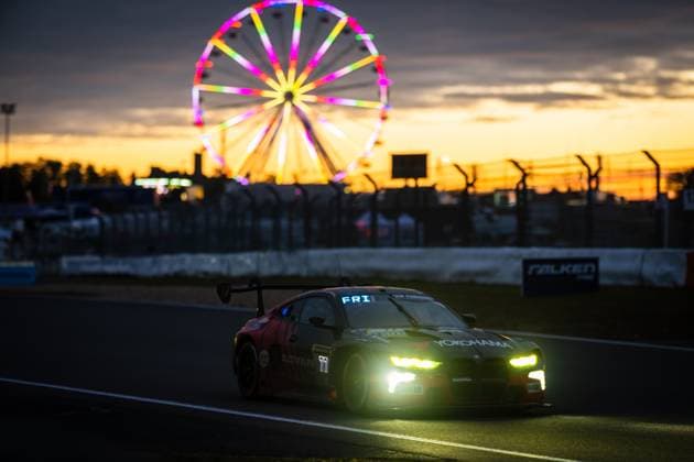 Der BMW M4 GT3 Charles Weerts & Marco Wittmann & Philipp Eng & Robin Frijns (Schubert Motorsport) von Schubert Motorsport fährt bei einbrechender Dämmerung über die Rennstrecke des Nürburgrings. Die eingeschalteten Scheinwerfer leuchten hell in der Abenddämmerung, während im Hintergrund ein farbenprächtiges, beleuchtetes Riesenrad den goldenen Sonnenuntergangshimmel schmückt. Sponsoren wie Yokohama und ADVAN sind auf der dunklen Fahrzeugkarosserie deutlich erkennbar. Das Falken-Werbebanner an der Streckenbegrenzung unterstreicht die Atmosphäre des 24-Stunden-Rennens. BMW M4 GT3 77 von Schubert Motorsport im Sonnenuntergang am Nürburgring Copyright: xMichaelxGelhardtx