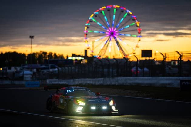 Der Mercedes-AMG GT3 Rennfahrzeug von Goroyan RT by Car Collection fährt in der Abenddämmerung über den Nürburgring. Die eingeschalteten Scheinwerfer leuchten intensiv, während im Hintergrund ein bunt beleuchtetes Riesenrad vor einem orangefarbenen Sonnenuntergangshimmel erstrahlt. Das Fahrzeug trägt die Red Bull-Lackierung mit auffälligem Artwork. Die stimmungsvolle Atmosphäre unterstreicht den Charakter des ADAC RAVENOL 24h Nürburgrings. Mercedes-AMG GT3 4 von Goroyan RT bei Sonnenuntergang am Nürburgring Copyright: xMichaelxGelhardtx