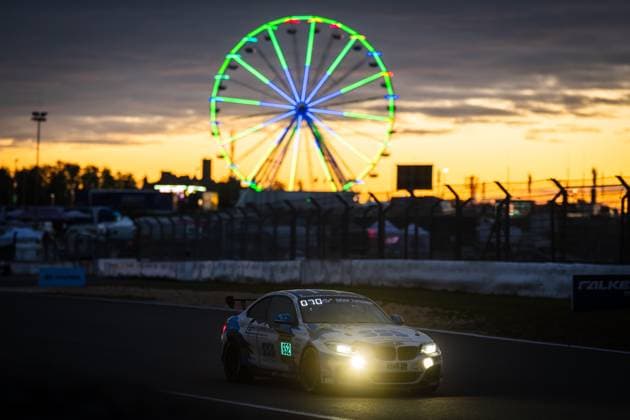 Der BMW M240i Racing Aldrin Opran & Axel Soyez & Grégoire Boutonnet & Lars Harbeck (Adrenalin Motorsport Team Mainhattan Wheels) von Adrenalin Motorsport Team Mainhattan Wheels fährt in der Abenddämmerung auf der Grand-Prix-Strecke des Nürburgrings. Die eingeschalteten Scheinwerfer erhellen die Fahrbahn, während im Hintergrund ein beleuchtetes Riesenrad vor einem orangefarbenen Sonnenuntergangshimmel leuchtet. Das Falken-Banner und die Boxenmauer sind im Mittelgrund zu erkennen. BMW 652 von Adrenalin Motorsport bei Sonnenuntergang am Nürburgring Copyright: xMichaelxGelhardtx