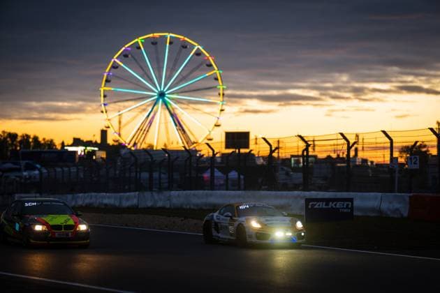 Zwei Fahrzeuge durchqueren bei beeindruckendem Sonnenuntergang die Strecke am Nürburgring. Im Vordergrund fährt ein weißer Porsche Cayman mit der Nummer 104, dahinter ein gelb-roter BMW. Im Hintergrund leuchtet das farbenfrohe Riesenrad des Fahrerlagers vor dem orange-violetten Abendhimmel. Ein Falken-Tyres-Banner ist an der Streckenbegrenzung zu sehen, die Scheinwerfer der Fahrzeuge erhellen die Dämmerung. Dramatische Sonnenuntergangs-Atmosphäre am Nürburgring mit Riesenrad Copyright: xMichaelxGelhardtx
