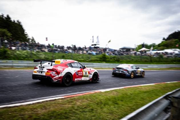 Der Toyota GR Supra Hugo Schwarze & Javier Sagrera (Toyota Racing United) von Toyota Racing United nimmt in einer schnellen Kurve am Nürburgring einen Gegner unter Druck. Das rot-weiße Fahrzeug mit den Sponsoren eFuel Group und Motul ist leicht versetzt und zeigt deutliche Bewegungsunschärfe, während im Hintergrund ein weiteres Fahrzeug in grau-blauer Lackierung folgt. Die Tribünen sind mit zahlreichen Zuschauern gefüllt. Toyota GR Supra 90 von Toyota Racing United im Duell am Nürburgring Copyright: xMichaelxGelhardtx