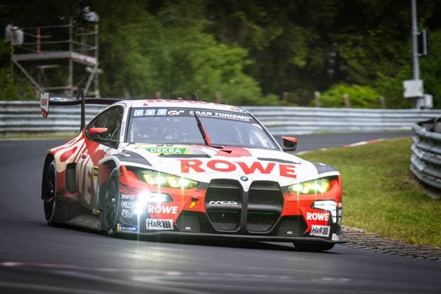 Der BMW M4 GT3 Dan Harper & Dries Vanthoor & Max Hesse & Sheldon van der Linde (ROWE RACING) von ROWE RACING nimmt eine Kurve auf der Nordschleife des Nürburgrings. Das Fahrzeug in der charakteristischen rot-weißen ROWE-Lackierung hat die Scheinwerfer eingeschaltet und zeigt eine dynamische Fahrhaltung. Sponsoren wie DEKRA, H&R und Michelin sind gut sichtbar auf der Karosserie. BMW M4 GT3 99 von ROWE RACING auf der Nordschleife Copyright: xMichaelxGelhardtx