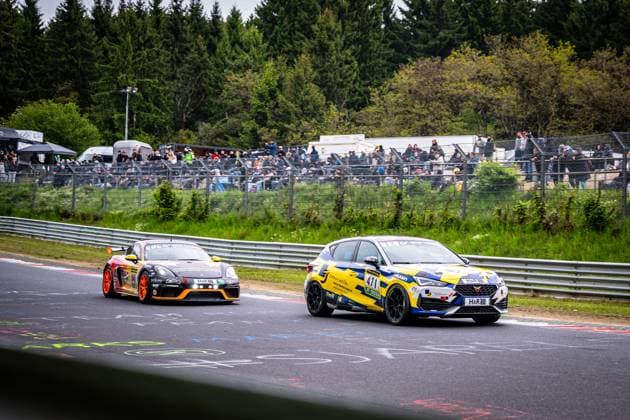 Zwei Fahrzeuge liefern sich ein enges Duell auf dem Nürburgring beim ADAC RAVENOL 24h Nürburgring 2026. Im Vordergrund fährt der Andreas Winterwerber & Michael Eichhorn & Tony Roma (Jung Motorsport) – ein CUPRA Leon Competición in gelb-blauem Design mit H&R- und Falken-Sponsoring – die Strecke entlang. Dahinter folgt ein schwarz-orangener Porsche 718 Cayman GT4 mit der Startnummer 441 und H&R-Sponsoring. Zahlreiche Zuschauer verfolgen das Rennen von der Tribüne hinter der Leitplanke, umgeben von der typischen grünen Eifel-Landschaft. CUPRA 471 und Porsche 441 kämpfen auf dem Nürburgring Copyright: xMichaelxGelhardtx