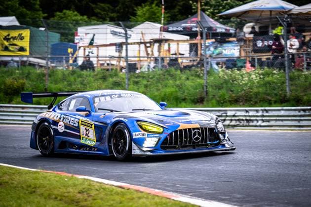 Der Mercedes-AMG GT3 Andreas Gülden & Tim Sandtler & Yuichi Nakayama (Toyo Tires with Ring Racing) von Toyo Tires with Ring Racing durchfährt eine Kurve auf der Nordschleife des Nürburgrings. Das blau-goldene Fahrzeug mit den markanten Toyo-Tires- und RAVENOL-Logos trägt sichtbare Unfallschäden an der Frontpartie – Karosserieteile sind mit Klebeband notdürftig gesichert. Die gelben Scheinwerfer leuchten, im Hintergrund sind Zuschauer und Zeltaufbauten zu erkennen. Mercedes-AMG GT3 32 von Toyo Tires with Ring Racing auf der Nordschleife Copyright: xMichaelxGelhardtx