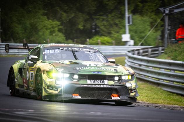 Der Ford Mustang GT3 Arjun Maini & Fabio Scherer & Frank Stippler (HRT Ford Racing) von HRT Ford Racing durchfährt mit eingeschalteten Scheinwerfern eine Kurve auf der Nordschleife am Nürburgring. Das Fahrzeug trägt ein markantes Camouflage-Lackierung in Grün- und Brauntönen mit dem Bundeswehr-Sponsoring. Sichtbare Sponsoren sind unter anderem H&R, Yokohama, RAVENOL und Sparco. Im Hintergrund sind die typischen Leitplanken und das üppige Grün der Eifel zu erkennen. Ford Mustang GT3 67 von HRT Ford Racing auf der Nordschleife Copyright: xMichaelxGelhardtx