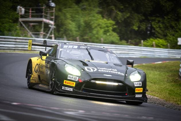 Der Aston Martin Vantage GT3 Christian Krognes & Mattia Drudi & Nicki Thiim & Felipe Fernandez Laser (natural elements by Walkenhorst Motorsport) von natural elements by Walkenhorst Motorsport durchfährt eine enge Kurve auf der Nordschleife des Nürburgrings. Das Fahrzeug in charakteristischem Dunkelgrün mit gelben Akzenten hat die Scheinwerfer eingeschaltet. Sponsoren wie natural elements, H&R und RAVENOL sowie Pirelli-Logos sind deutlich sichtbar. Im Hintergrund sind Leitplanken, ein Streckenposten-Turm und dichter Baumbestand zu erkennen. Aston Martin GT3 34 von natural elements by Walkenhorst Motorsport auf der Nordschleife Copyright: xMichaelxGelhardtx