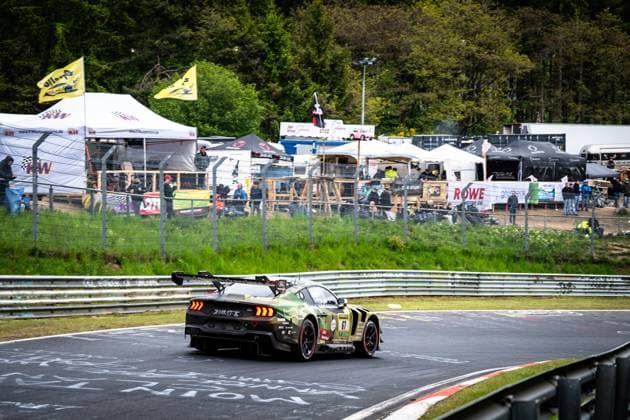 Der Ford Mustang GT3 von HRT Ford Racing durchfährt eine Kurve auf der Nordschleife am Nürburgring. Das Fahrzeug Arjun Maini & Fabio Scherer & Frank Stippler (HRT Ford Racing) trägt ein markantes Tarnmuster-Lackierung in Oliv- und Schwarztönen und ist von hinten zu sehen, wobei der große Heckflügel und die leuchtenden Bremslichter gut erkennbar sind. Im Hintergrund sind zahlreiche Zuschauer, Teamzelte sowie Werbebanner – darunter KW und ROWE Motor Oil – zu sehen. Ford Mustang GT3 67 von HRT Ford Racing auf der Nordschleife Copyright: xMichaelxGelhardtx