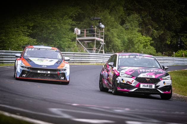 Zwei Fahrzeuge liefern sich auf der Nordschleife des Nürburgrings einen engen Zweikampf. Links kämpft ein Hyundai Elantra N TCR in blau-orangem Design mit H&R-Sponsoring und Gran Turismo-Branding um Position. Rechts fährt ein CUPRA Leon in einem auffälligen pink-schwarz-weißen Camouflage-Design, ebenfalls mit H&R-Sponsoring und Gran Turismo-Branding. Beide Fahrzeuge durchfahren eine schnelle Kurve auf dem grünen Waldabschnitt der Nordschleife. Die Startnummern beider Fahrzeuge sind aufgrund der Fahrt und des Bildwinkels nicht eindeutig lesbar. Hyundai und CUPRA im Zweikampf auf der Nordschleife Copyright: xMichaelxGelhardtx