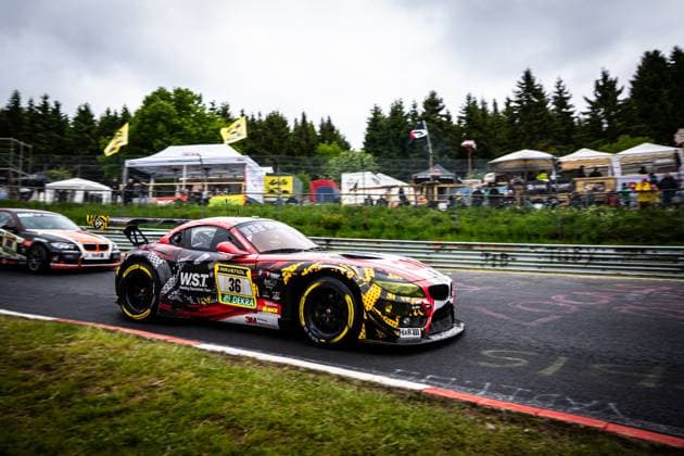 Der BMW Z4 GT3 Christian Scherer & Henry Walkenhorst & Julian Reeh & Ralf Schall (Saugmotoren Motorsport 3M) von Saugmotoren Motorsport 3M nimmt eine Kurve auf der Nordschleife des Nürburgrings. Das Fahrzeug trägt ein auffälliges rot-schwarz-gelbes Camouflage-Design mit Sponsoren wie W.S.T. Welding Specialists Team, RAVENOL, 3M und DEKRA. Im Hintergrund sind Zuschauer, Zelte und die typische Eifel-Landschaft mit dichten Nadelwäldern zu sehen. Ein weiteres Fahrzeug ist im Hintergrund zu erkennen. BMW Z4 GT3 36 von Saugmotoren Motorsport 3M auf der Nordschleife Copyright: xMichaelxGelhardtx