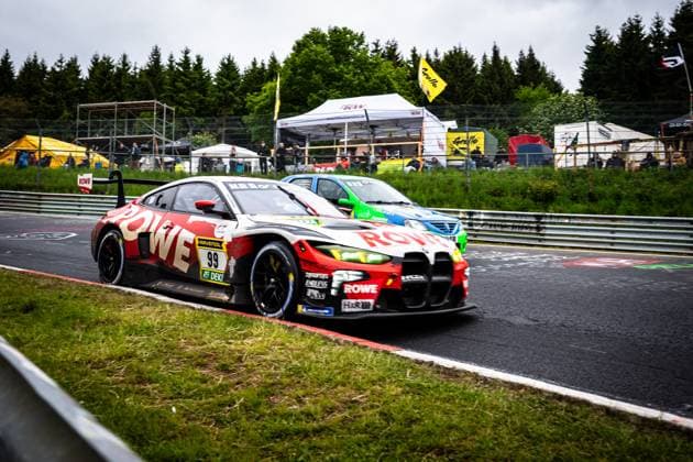 Der Dan Harper & Dries Vanthoor & Max Hesse & Sheldon van der Linde (ROWE RACING) von ROWE RACING durchfährt eine Kurve auf der Nordschleife am Nürburgring. Das Fahrzeug in der markanten rot-weißen ROWE-Lackierung trägt die Startnummer 99 und zeigt Sponsoren wie RAVENOL, ENDLESS und H&R. Im Hintergrund ist ein weiteres Fahrzeug in blau-grüner Lackierung zu sehen, während zahlreiche Zuschauer das Geschehen am Streckenrand verfolgen. BMW M4 GT3 99 von ROWE RACING auf der Nordschleife Copyright: xMichaelxGelhardtx