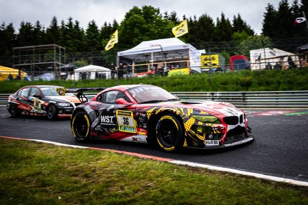 Der BMW Z4 GT3 Christian Scherer & Henry Walkenhorst & Julian Reeh & Ralf Schall (Saugmotoren Motorsport 3M) von Saugmotoren Motorsport 3M nimmt eine Kurve auf der Nordschleife am Nürburgring. Das Fahrzeug trägt ein auffälliges rot-schwarz-gelbes Livery mit Sponsoren wie W.S.T. Welding Specialists Team, RAVENOL, DEKRA und 3M. Im Hintergrund folgt ein weiteres Fahrzeug mit der Startnummer 100, während Zuschauer und Teamzelte die Strecke säumen. BMW Z4 GT3 36 von Saugmotoren Motorsport 3M auf der Nordschleife Copyright: xMichaelxGelhardtx