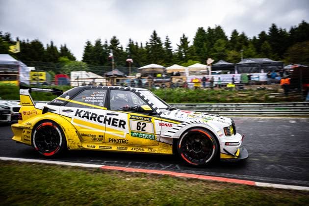 Der Luca Ludwig & Markus Winkelhock & Sebastian Asch (HWA Engineering Speed) von HWA Engineering Speed durchfährt mit hohem Tempo eine Kurve auf der Nordschleife am Nürburgring. Das markante gelb-weiß-schwarze Fahrzeug trägt prominente Sponsorenlogos wie Kärcher, RAVENOL, SONAX und bluechem GROUP. Im nassen Asphalt spiegelt sich die bedeckte Witterung der Eifel, im Hintergrund sind Zuschauer und Hospitality-Zelte sichtbar. Mercedes-AMG 62 von HWA Engineering Speed auf der Nordschleife Copyright: xMichaelxGelhardtx