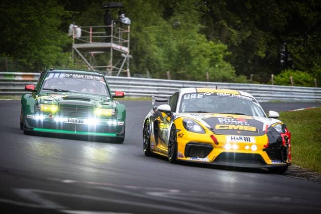 Zwei Fahrzeuge kämpfen im Duell auf der nassen Nordschleife des Nürburgrings. Im Vordergrund fährt ein gelb-schwarzer Porsche 718 Cayman GT4 mit CMB- und H&R-Sponsoring, während dahinter ein dunkelgrüner Rennwagen mit PARKSIDE- und H&R-Sponsoring sowie Gran-Turismo-Branding dicht aufsetzt. Die Fahrbahn ist feucht, im Hintergrund ist grüner Waldrand mit einem Streckenposten-Gerüst zu erkennen. Porsche 718 Cayman GT4 und grüner Rennwagen auf der Nordschleife Copyright: xMichaelxGelhardtx