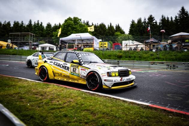 Der Mercedes-Benz 190E Evolution von HWA Engineering Speed mit der Startnummer 62 durchfährt eine Kurve auf der Nordschleife des Nürburgrings. Das markante gelb-weiße Fahrzeug mit KÄRCHER- und RAVENOL-Sponsoring präsentiert sich in einem klassischen Rennwagen-Design mit modernem Aerodynamik-Paket. Im Hintergrund ist ein weiteres Fahrzeug sowie das Fahrerlager mit Zuschauern und Zelten zu erkennen. Das Rennen findet bei bewölktem Wetter statt. Der Platzhalter Luca Ludwig & Markus Winkelhock & Sebastian Asch (HWA Engineering Speed) kennzeichnet dieses Fahrzeug. Mercedes-Benz 190E 62 von HWA Engineering Speed auf der Nordschleife Copyright: xMichaelxGelhardtx