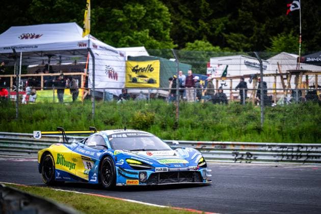 Der McLaren GT3 Ben Doerr & Marvin Kirchhöfer & Timo Glock & Timo Scheider (Doerr Motorsport) von Doerr Motorsport durchfährt eine enge Rechtskurve auf der Nürburgring-Nordschleife. Das Fahrzeug trägt die auffällige blau-gelbe Lackierung mit dem Bitburger-Sponsoring und der Startnummer 69. Im Hintergrund sind Zuschauer und die Zelte der Teams sichtbar, darunter ein KW-Suspensions-Banner sowie das Grello-Logo. McLaren 69 von Doerr Motorsport in der Kurve am Nürburgring Copyright: xMichaelxGelhardtx