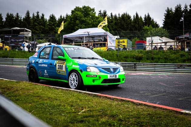Der Dacia Logan Robert Neumann & Alexander Becker & Christian Geilfus & Gregor Starck & Oliver Kriese (Ollis Garage Racing) von Ollis Garage Racing durchfährt eine Kurve auf dem Nürburgring. Das Fahrzeug ist in einem auffälligen Blau-Grün-Farbschema lackiert und trägt Sponsoren wie RAVENOL, DEKRA, Falken, Goodyear, junited AUTOGLAS, H&R und Bleker Autoteile. Im Hintergrund sind Zuschauer, gelbe Fahnen und ein weißes Zelt zu sehen. Dacia Logan 300 von Ollis Garage Racing auf dem Nürburgring Copyright: xMichaelxGelhardtx