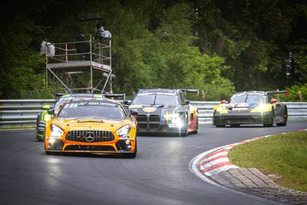 Der orange Mercedes-AMG GT3 von Mercedes-AMG Team RAVENOL (Fabian Schiller & Luca Stolz & Maro Engel & Maxime Martin (Mercedes-AMG Team RAVENOL)) führt ein Gruppe von Fahrzeugen durch eine Kurve auf der Nordschleife am Nürburgring. Mit eingeschalteten Scheinwerfern und dem markanten RAVENOL-Sponsoring nimmt das Fahrzeug den Scheitelpunkt der Kurve. Im Hintergrund folgen ein BMW M4 GT3 sowie ein weiteres Fahrzeug – beide mit eingeschalteten Lichtern. Links im Bild ist zudem ein grünes Fahrzeug zu erkennen. Eine Kamerabrücke mit Fotografen ist im Hintergrund aufgebaut. Mercedes-AMG GT3 80 von Mercedes-AMG Team RAVENOL auf der Nordschleife Copyright: xMichaelxGelhardtx