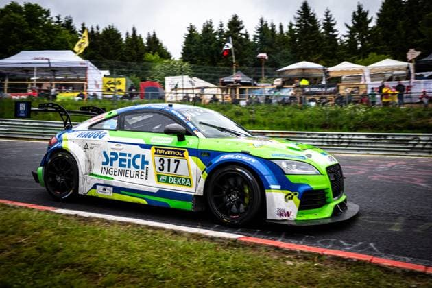 Der Audi TT RS von 2R Racing passiert mit der Startnummer 317 einen Streckenabschnitt auf der Nordschleife am Nürburgring. Das Fahrzeug Roland Waschkau & Dirk Vleugels & Thorsten Jung & Wolfgang Haugg (2R Racing) trägt ein auffälliges grün-blau-weißes Lackdesign mit den Sponsoren Speich Ingenieure GmbH, RAVENOL und Falken. Im Hintergrund sind Zuschauer und Hospitality-Zelte der Teamlager zu erkennen, während das Fahrzeug mit Bewegungsunschärfe durch die Kurve fährt. Audi TT 317 von 2R Racing auf der Nordschleife Copyright: xMichaelxGelhardtx