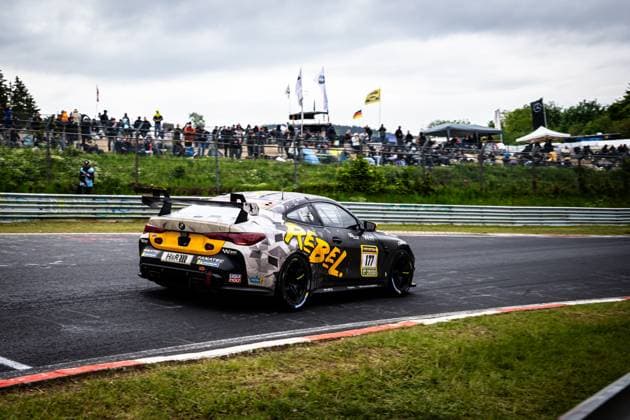Der BMW M4 GT4 Alexandru Vasilescu & David Ogburn & Malcolm Harrison & Mark Smith & Sergiu Nicolae (AV Racing by BLACK FALCON) von AV Racing by BLACK FALCON durchfährt eine Kurve auf dem Nürburgring. Das Fahrzeug trägt ein markantes Camouflage-Design in Grau und Schwarz mit auffälligen gelben Akzenten und dem Schriftzug REBEL . Zahlreiche Sponsoren wie LIQUI MOLY, Fanatec und H&R sind auf der Karosserie sichtbar. Im Hintergrund verfolgen zahlreiche Zuschauer das Renngeschehen beim ADAC RAVENOL 24h Nürburgring. BMW M4 GT4 177 von AV Racing by BLACK FALCON auf dem Nürburgring Copyright: xMichaelxGelhardtx
