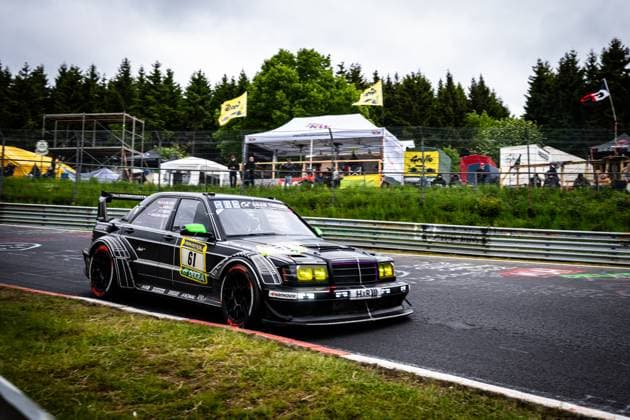 Der markante schwarze Mercedes 190E mit der Startnummer 61 von HWA Engineering Speed nimmt eine Kurve auf der Nordschleife des Nürburgrings. Das Fahrzeug präsentiert sich mit breitem Aeropaket, gelben Scheinwerfern und auffälligen roten Reifenseitenwänden. Sponsoren wie RAVENOL, DEKRA und H&R sind deutlich sichtbar. Im Hintergrund verfolgen Zuschauer das Rennen unter bedecktem Himmel. Bruno Spengler & Christian Gebhardt & Evald Holstad & Peter Ludwig (HWA Engineering Speed) Mercedes 190E 61 von HWA Engineering Speed auf der Nordschleife Copyright: xMichaelxGelhardtx