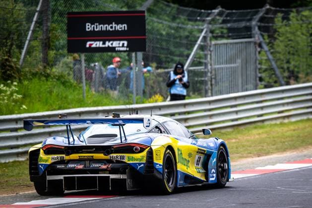 Der McLaren 720S GT3 mit der Startnummer 69 von Doerr Motorsport passiert in Blau-Gelb den ikonischen Streckenabschnitt Brünnchen auf der Nürburgring-Nordschleife. Das Fahrzeug trägt die Sponsoren Bitburger, RAVENOL und H&R prominent auf der Karosserie. Im Hintergrund sind Zuschauer und ein Streckenposten zu sehen, dahinter das charakteristische Brünnchen-Schild mit Falken Tyres-Branding. Ben Doerr & Marvin Kirchhöfer & Timo Glock & Timo Scheider (Doerr Motorsport) McLaren 69 von Doerr Motorsport am Brünnchen auf der Nordschleife Copyright: xMichaelxGelhardtx
