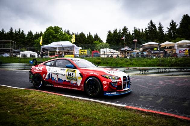 Der BMW M4 GT4 Aris Balanian & Jeroen Bleekemolen & Joshua Bednarski & Peter Cate & Tom Schütze (Riller & Schnauck powered by Cerny Motorsport) von Riller & Schnauck powered by Cerny Motorsport durchfährt einen Kurvenabschnitt auf der Nordschleife des Nürburgrings. Das Fahrzeug trägt ein markantes rot-weißes Stadtsilhouetten-Design mit den Sponsoren NGK Spark Plugs, RAVENOL und Falken sowie dem DEKRA-Logo. Im Hintergrund sind Zuschauerbereiche und Zelte unter bedecktem Himmel zu erkennen. BMW M4 GT4 145 von Riller & Schnauck powered by Cerny Motorsport auf der Nordschleife Copyright: xMichaelxGelhardtx