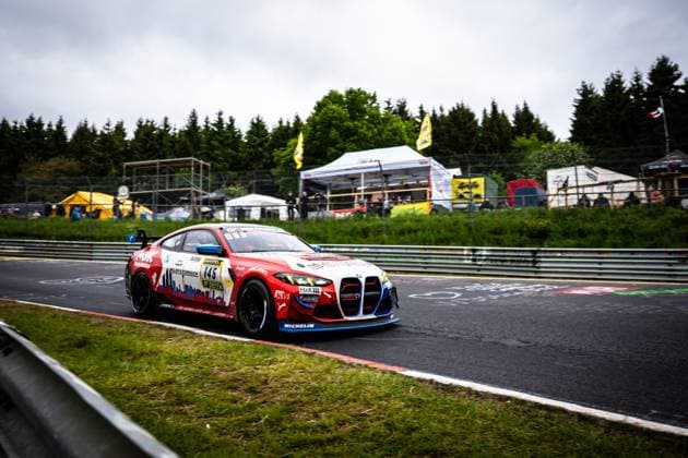 Der Aris Balanian & Jeroen Bleekemolen & Joshua Bednarski & Peter Cate & Tom Schütze (Riller & Schnauck powered by Cerny Motorsport) von Riller & Schnauck powered by Cerny Motorsport durchfährt eine Kurve auf dem Nürburgring. Das rot-weiße BMW M4 GT4 mit der Startnummer 145 ist mit Sponsoren wie NGK, Michelin, H&R und DEKRA beklebt. Im Hintergrund sind Zuschauer, Zelte und gelbe Fahnen zu sehen. BMW M4 GT4 145 von Riller & Schnauck powered by Cerny Motorsport auf der Nordschleife Copyright: xMichaelxGelhardtx