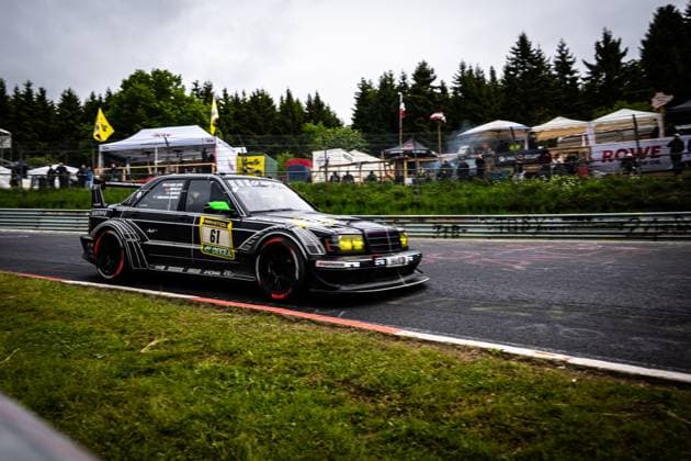 Der schwarz lackierte Mercedes-Benz Rennwagen Bruno Spengler & Christian Gebhardt & Evald Holstad & Peter Ludwig (HWA Engineering Speed) von HWA Engineering Speed durchfährt eine Kurve auf dem Nürburgring. Das markant gestaltete Fahrzeug mit gelben Scheinwerfern und auffälligem Aerodynamikpaket trägt die Startnummer 61 mit RAVENOL- und DEKRA-Sponsoring. Im Hintergrund sind Zuschauer, Zelte und das ROWE-Banner des Fahrerlagers zu erkennen. Mercedes-Benz 61 von HWA Engineering Speed auf dem Nürburgring Copyright: xMichaelxGelhardtx