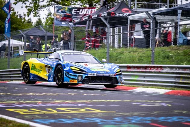 Der McLaren GT3 Ben Doerr & Marvin Kirchhöfer & Timo Glock & Timo Scheider (Doerr Motorsport) von Doerr Motorsport durchfährt eine Kurve auf der Nordschleife des Nürburgrings. Das Fahrzeug trägt das auffällige blau-gelbe Bitburger-Livery und ist mit LED-Scheinwerfern ausgestattet. Sponsoren wie H&R und Pirelli sind deutlich sichtbar. Im Hintergrund verfolgen zahlreiche Zuschauer das Rennen. McLaren GT3 69 von Doerr Motorsport auf dem Nürburgring Copyright: xMichaelxGelhardtx