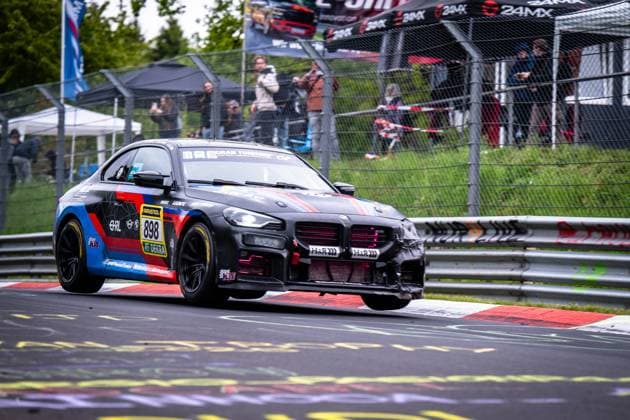 Der BMW M2 898; Frauenfußball Nationalmannschaft Finnland von Hofor Racing by Bonk Motorsport nimmt eine Kurve auf der Nordschleife des Nürburgrings beim ADAC RAVENOL 24h Nürburgring 2026. Das schwarz-blau-rote Fahrzeug mit den Sponsoren RAVENOL, H&R und DEKRA hebt auf dem charakteristischen rot-weißen Curb leicht ab. Im Hintergrund verfolgen zahlreiche Zuschauer das Renngeschehen hinter dem Fangzaun. BMW M2 898 von Hofor Racing by Bonk Motorsport auf der Nordschleife Copyright: xMichaelxGelhardtx