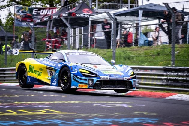 Der McLaren GT3 Ben Doerr & Marvin Kirchhöfer & Timo Glock & Timo Scheider (Doerr Motorsport) von Doerr Motorsport nimmt mit gesenkter Vorderachse dynamisch einen Curb auf der Nordschleife am Nürburgring. Das Fahrzeug trägt das auffällige blau-gelbe Bitburger-Livery mit H&R- und Pirelli-Sponsoring. Im Hintergrund verfolgen zahlreiche Zuschauer das Rennen vom Streckenrand aus. McLaren GT3 69 von Doerr Motorsport auf der Nordschleife Copyright: xMichaelxGelhardtx