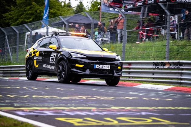 Ein schwarz-gelbes Cupra Race Management Fahrzeug (Race Management 3) fährt auf der Nordschleife des Nürburgrings. Das Fahrzeug ist mit einem orangefarbenen Blaulichtbalken auf dem Dach ausgestattet und trägt die Logos von RAVENOL, Löhr Gruppe und dem 24h Nürburgring. Im Hintergrund sind Streckenposten, Fans an der Absperrung sowie Werbebanner zu sehen. Race Management Fahrzeug auf der Nordschleife beim 24h Nürburgring Copyright: xMichaelxGelhardtx