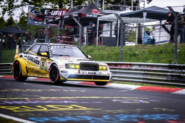 Der spektakuläre Mercedes-Benz 190E mit der Startnummer 62 von HWA Engineering Speed nimmt eine Kurve auf der Nordschleife des Nürburgrings mit eindrucksvoller Dynamik. Das in Gelb und Weiß gehaltene Fahrzeug trägt die Sponsoren Kärcher, RAVENOL, SONAX, H&R und Hankook und ist mit Gran-Turismo-Branding versehen. Im Hintergrund sind Zuschauer hinter dem Sicherheitszaun sowie ein 24MX-Zelt zu sehen. Die eingeschalteten Scheinwerfer und der charakteristische Heckspoiler unterstreichen den Renncharakter des historischen Fahrzeugs. Luca Ludwig & Markus Winkelhock & Sebastian Asch (HWA Engineering Speed) Mercedes-Benz 190E 62 von HWA Engineering Speed auf der Nordschleife Copyright: xMichaelxGelhardtx