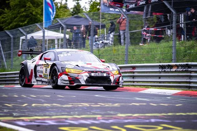 Der Audi R8 LMS GT3 Christian Kohlhaas & Duncan Huismann & Jan Jaap van Roon & Rudy van Buren (Max Kruse Racing) von Max Kruse Racing durchfährt eine Kurve auf der Nordschleife des Nürburgrings. Das Fahrzeug trägt die Startnummer 19 und zeigt ein markantes weiß-rot-graues Camouflage-Design mit Sponsoren wie MASCOT WORKWEAR, RAVENOL und H&R. Im Hintergrund verfolgen Zuschauer das Rennen hinter dem Fangzaun. Audi R8 LMS GT3 19 von Max Kruse Racing auf der Nordschleife Copyright: xMichaelxGelhardtx