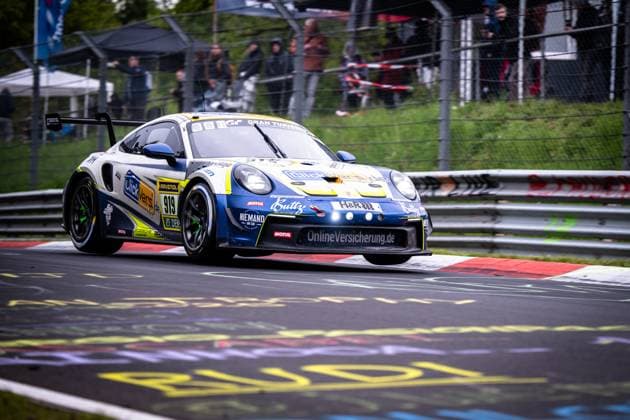 Der Porsche 911 GT3 von Clickversicherung.de TEAM (Kersten Jodexnis & Peter Scharmach & Richard Jodexnis & Richard-Sven Jodexnis & Robin Chrzanowski (Clickversicherung.de TEAM)) nimmt die roten Curbs des Nürburgrings in einer dynamischen Kurvenfahrt. Das in Weiß und Blau gehaltene Fahrzeug trägt Sponsoren wie RAVENOL, MOTUL und OnlineVersicherung.de und ist mit der Startnummer 919 klar erkennbar. Im Hintergrund verfolgen Zuschauer hinter den Zäunen das Renngeschehen. Porsche 911 GT3 919 von Clickversicherung.de TEAM auf der Nordschleife Copyright: xMichaelxGelhardtx