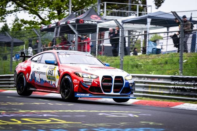 Der BMW M4 GT4 Aris Balanian & Jeroen Bleekemolen & Joshua Bednarski & Peter Cate & Tom Schütze (Riller & Schnauck powered by Cerny Motorsport) von Riller & Schnauck powered by Cerny Motorsport nimmt eine Kurve auf der Nordschleife des Nürburgrings. Das Fahrzeug in auffälligem Rot-Blau-Weiß-Design hebt leicht mit dem Vorderrad ab, was die dynamische Fahrsituation unterstreicht. Zu den sichtbaren Sponsoren zählen NGK Spark Plugs, H&R, Michelin und DEKRA. Im Hintergrund verfolgen Zuschauer das Geschehen hinter dem Fangzaun. BMW M4 GT4 145 von Riller & Schnauck powered by Cerny Motorsport am Nürburgring Copyright: xMichaelxGelhardtx