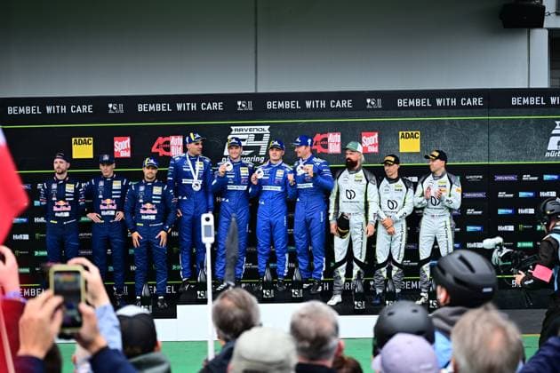 Siegerehrung auf dem Podium der 24h Nürburgring mit Fahrern in blauen und weißen Rennanzügen. Die Teams präsentieren ihre Pokale und Medaillen vor der Bembel with Care Bande und Sponsorenwand. Zuschauer fotografieren die Siegerfahrer im Fahrerlager. Siegerehrung 24h Nürburgring 2026 mit Fahrern und Teams auf dem Podium Copyright: xMichaelxGelhardtx