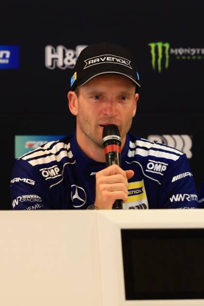 Maro Engel spricht bei einer Pressekonferenz im Fahrerlager des 24h Nürburgring. Der Fahrer der Mercedes-AMG Team RAVENOL trägt die charakteristische dunkelblaue Teamkleidung mit OMP und RAVENOL-Branding. Maro Engel bei Pressekonferenz der Mercedes-AMG Team RAVENOL