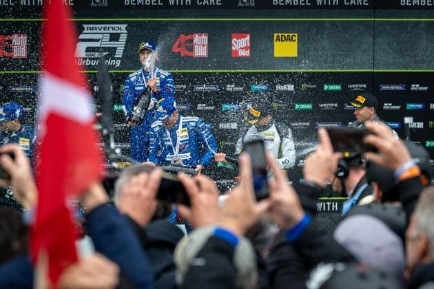 Christian Krognes steht auf dem Podium und wird mit Champagner bespritzt während Fotografen und Fans die Siegerehrung dokumentieren. Im Hintergrund sind Sponsor-Banner mit RAVENOL, ADAC und weiteren Partnern erkennbar. Siegerehrung auf dem Podium des 24h Nürburgring 2026 Copyright: xMichaelxGelhardtx