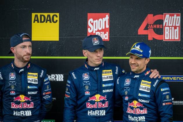 Drei Fahrer des Red Bull Team ABT präsentieren sich bei einer Pressekonferenz. Die Fahrer tragen die charakteristischen dunkelblauen Overalls mit Red Bull-, RAVENOL-, SONAX- und Michelin-Sponsoring sowie ABT-Branding. Im Hintergrund sind Medienlogos wie ADAC, Bild und Auto Bild zu sehen. Red Bull Team ABT Fahrer bei Pressekonferenz zur 24h Nürburgring 2026 Copyright: xMichaelxGelhardtx