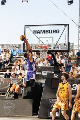 Beim FIBA 3x3 Hamburg Challenger in der Active City Arena zeigt ein Spieler in violettem Trikot einen beeindruckenden Dunking-Versuch zum Korb, während Spieler in gelben Trikots der Barcelona-Mannschaft die Aktion verfolgen. Das Event fand am 23. Mai 2026 in Hamburg statt. Spektakulärer Dunking-Moment beim FIBA 3x3 Hamburg Challenger
