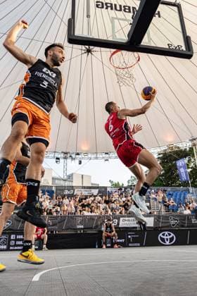 Spannendes 3x3-Basketball-Match beim FIBA 3x3 Hamburg Challenger am 23. Mai 2026 in der Active City Arena. Ein Spieler von Fuengirola (rotes Trikot) mit der Nummer 6 startet einen Korbleger, während ein Spieler von Amsterdam RABOBANK (schwarzes Trikot mit orangefarbenem Beinsport) mit erhobener Faust versucht, die Aktion zu verteidigen. Fuengirola ESP gegen Amsterdam RABOBANK NED – 3x3 Basketball in Hamburg